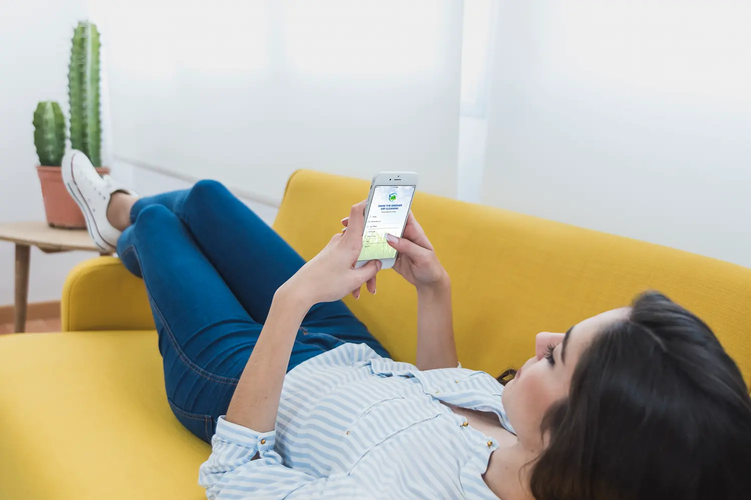 A woman lies on a yellow couch, holding and looking at her smartphone. A potted cactus sits on a small table nearby.