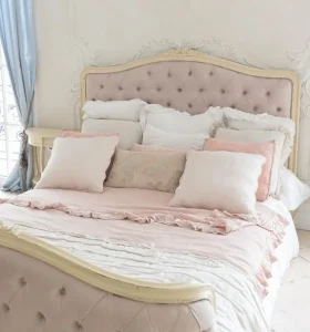 A neatly made bed with a tufted headboard, covered in pastel pink and white bedding, multiple pillows, and a light blue curtain on the left.