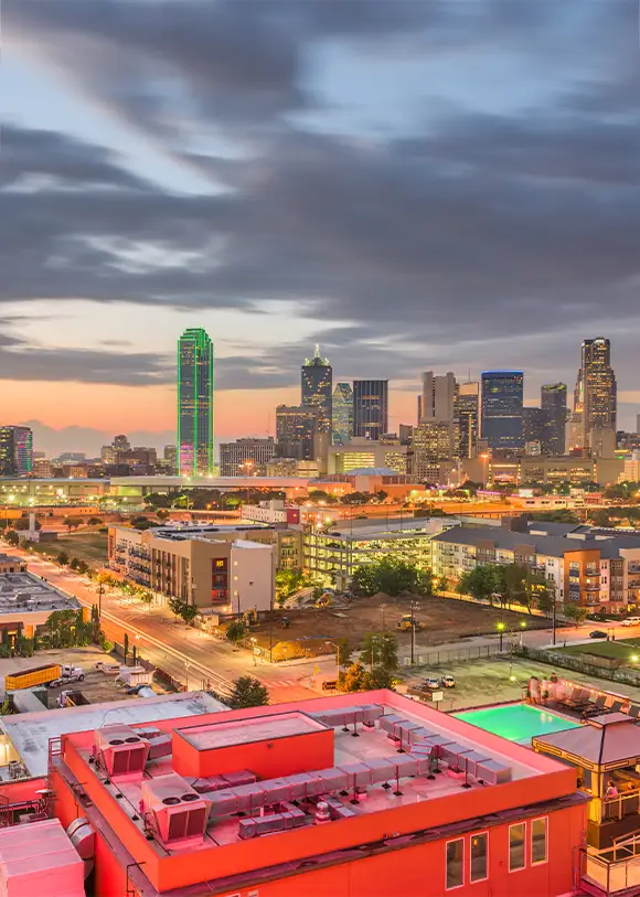 A city skyline at dusk with lit buildings, including a tall tower with green lights, under a partly cloudy sky. Foreground features red rooftops and urban streets.