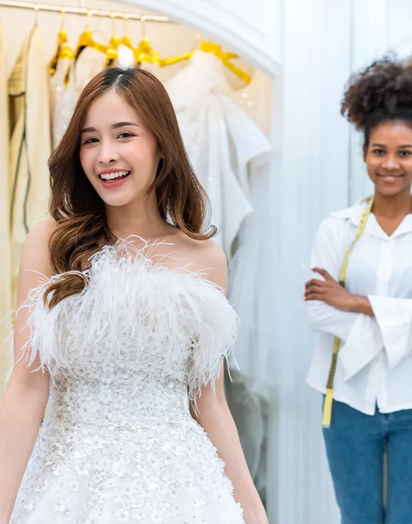 A woman in a feathered white wedding dress smiles, while another woman with a measuring tape around her neck stands in the background, arms crossed.