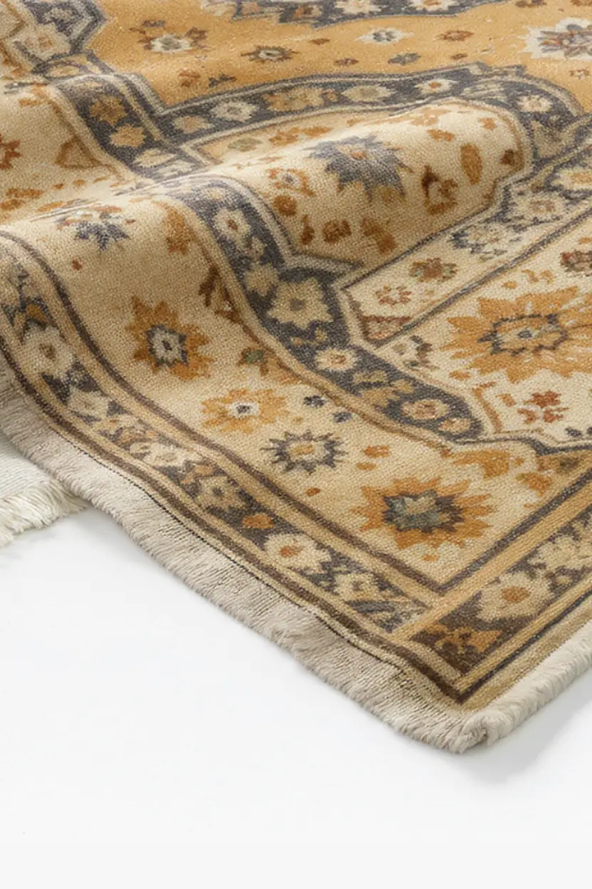 A beige and gold patterned area rug with floral motifs and fringed edges is shown partially folded on a white surface.