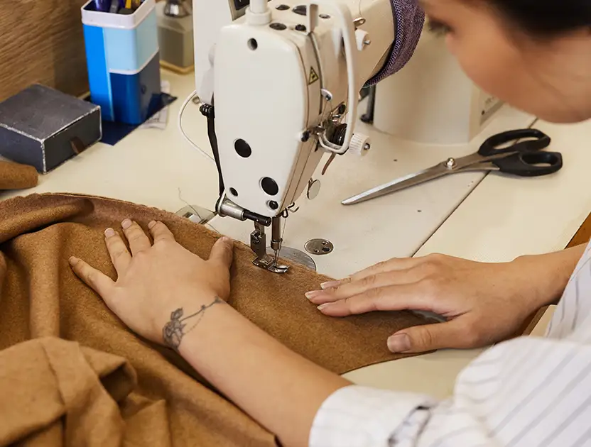 Person using a sewing machine to stitch brown fabric, with a pair of scissors and sewing supplies nearby on the worktable.