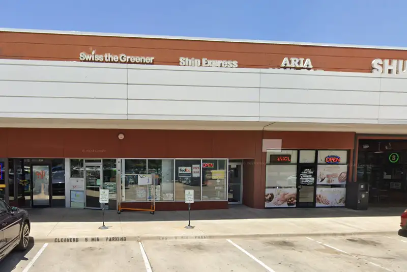 A row of storefronts in a shopping center includes Swiss the Greener cleaners, Ship Express, and a nail salon, with parking spaces in front and signs above each business.