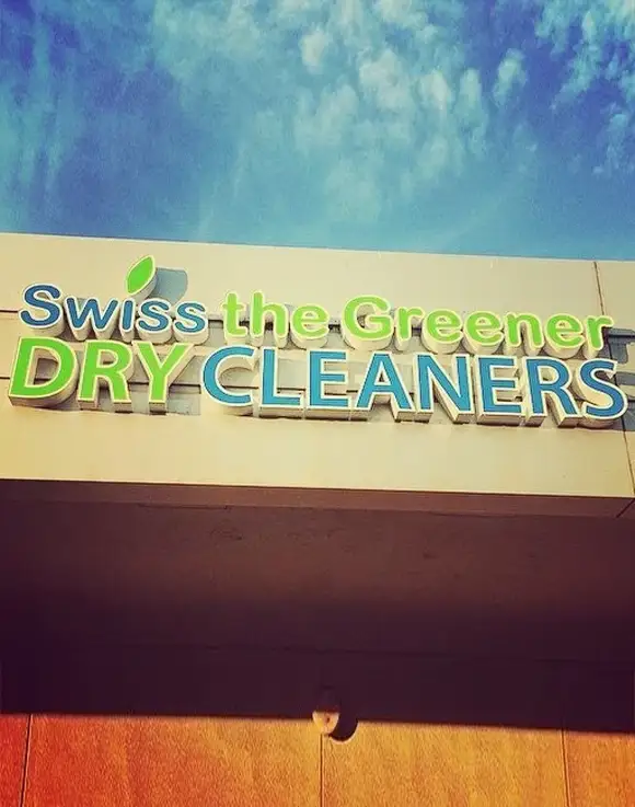 Sign for "Swiss the Greener DRY CLEANERS" with green and blue text against a white background, mounted on a building under a partly cloudy sky.