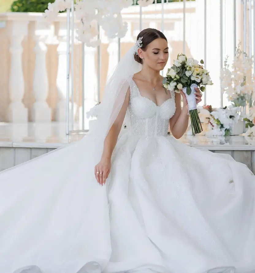 A bride in a white wedding gown and veil sits holding a bouquet of flowers, posing indoors with white floral decorations in the background.