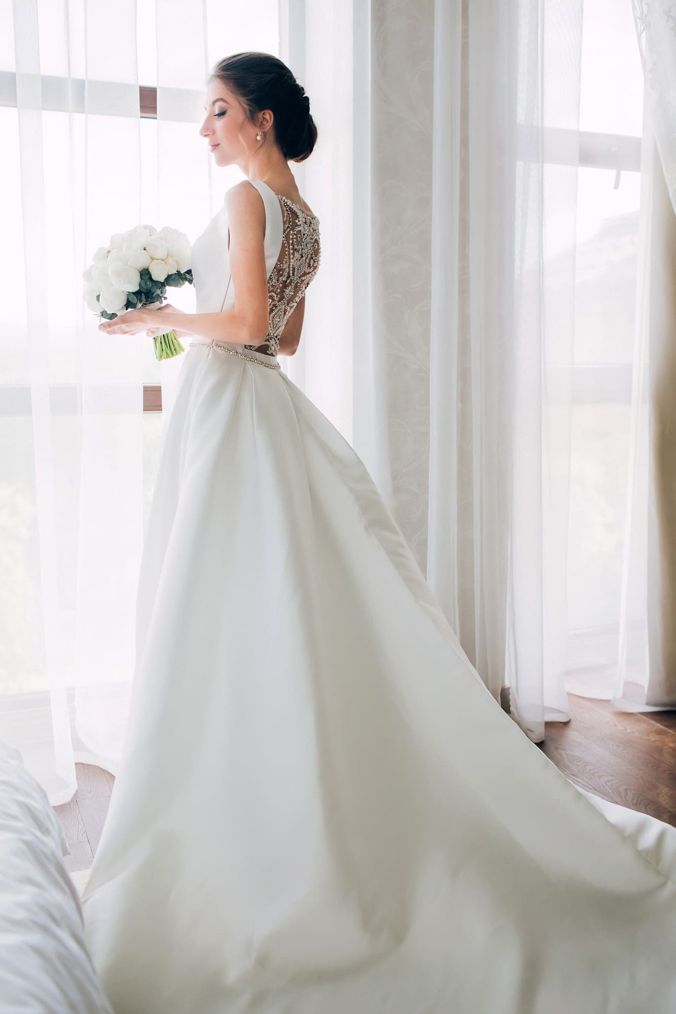 A bride in a white wedding dress holding a bouquet of white flowers stands by a window with sheer curtains, looking outside.