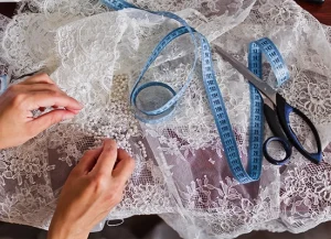 Hands sewing beads onto white lace fabric, with a blue measuring tape and a pair of scissors lying nearby on the table.
