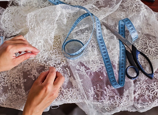 Hands sewing beads onto white lace fabric, with a blue measuring tape and a pair of scissors lying nearby on the table.