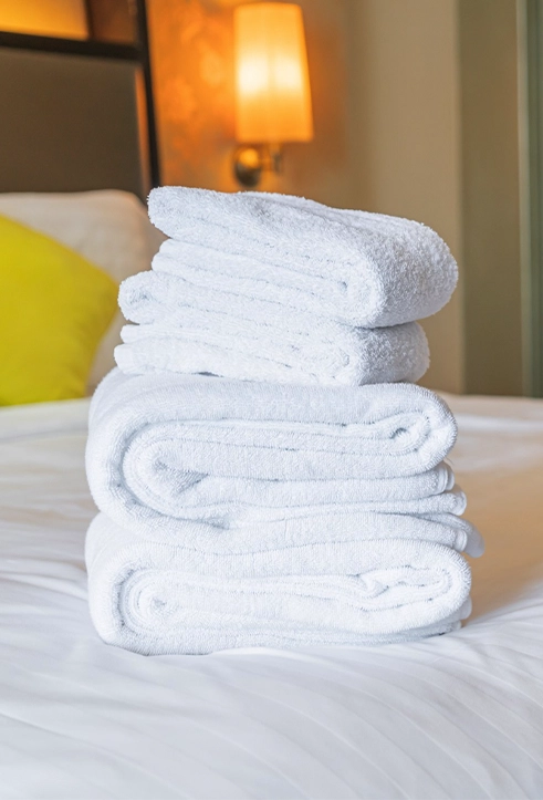 A neatly stacked pile of white towels sits on a made bed in a well-lit room with a lamp and a yellow cushion in the background.