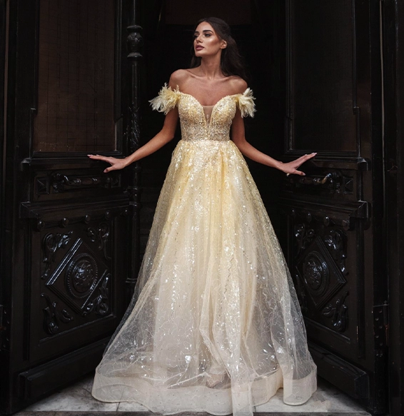 Woman wearing an off-shoulder, yellow-gold, sequined ball gown stands in an ornate doorway with dark wooden doors.