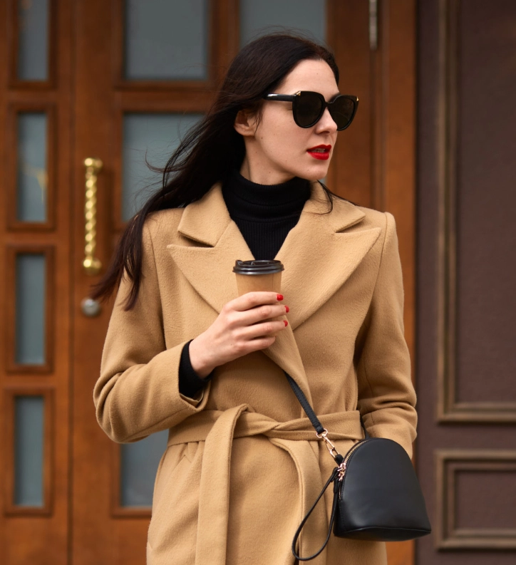 A woman in a tan coat and black sunglasses holds a coffee cup and a black handbag while standing in front of wooden doors.
