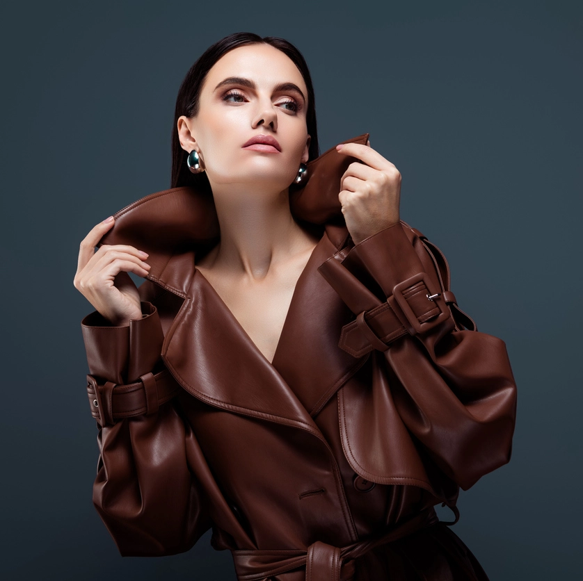 A woman in a brown leather trench coat poses with the collar raised, looking upward against a plain dark background.