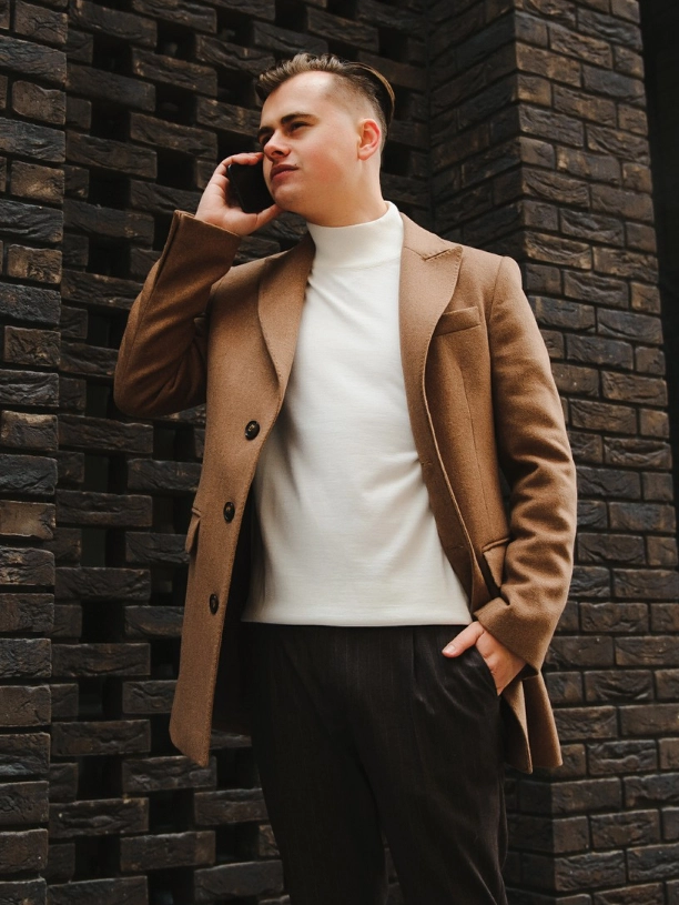 A man in a brown coat and white turtleneck is standing outdoors by a brick wall, holding a phone to his ear and looking to the side.