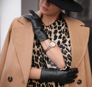 A woman wearing a camel coat, leopard print dress, black leather gloves, a gold wristwatch, and a black hat stands with her arms crossed.