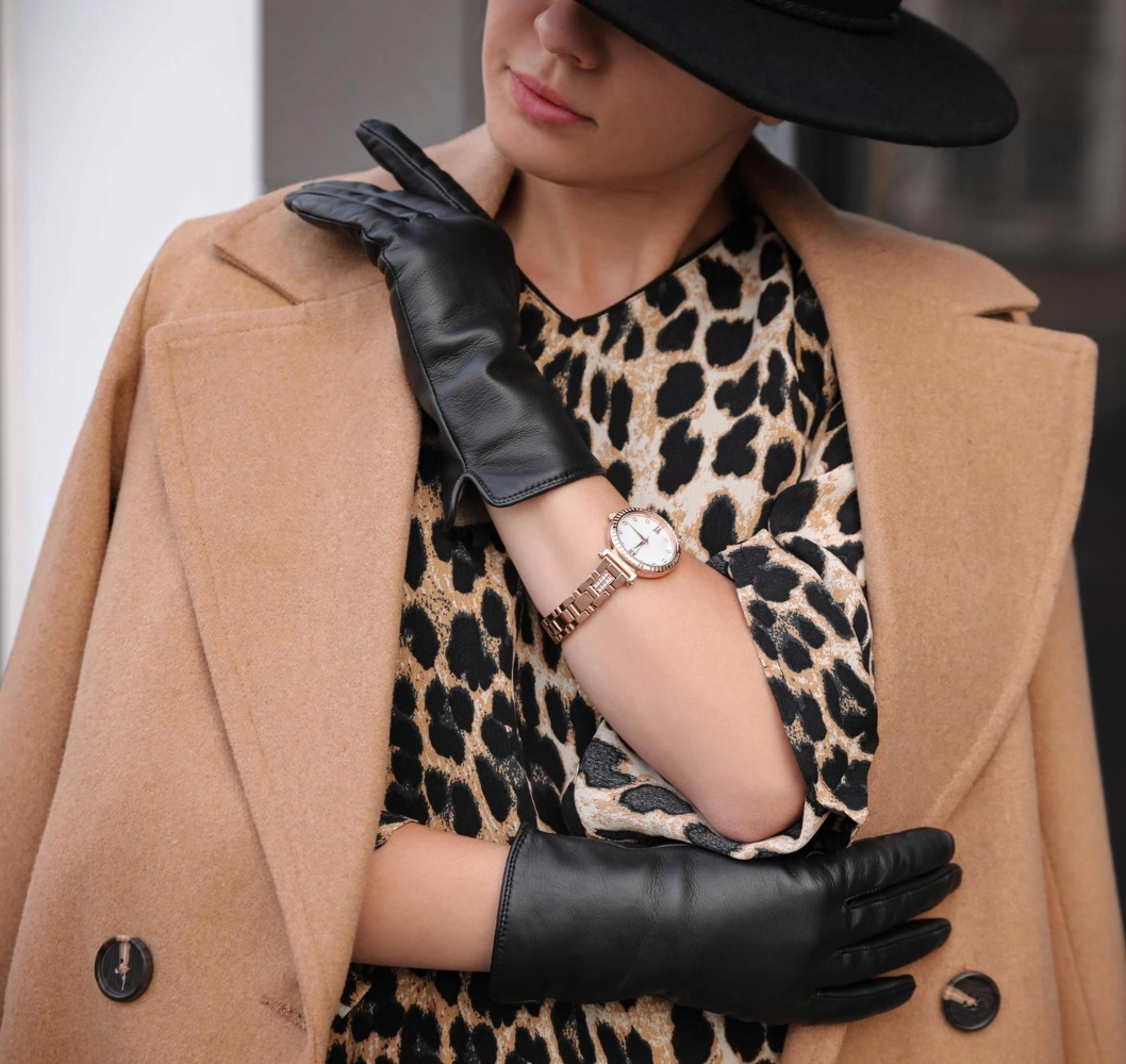 A woman wearing a camel coat, leopard print dress, black leather gloves, a gold wristwatch, and a black hat stands with her arms crossed.