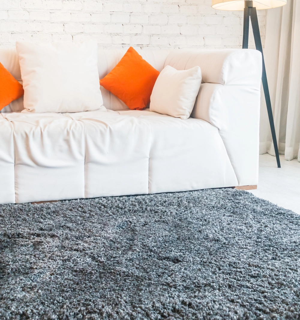 A modern living room with a white sofa, white and orange cushions, a gray shag rug, a white brick wall, and a floor lamp in the corner.