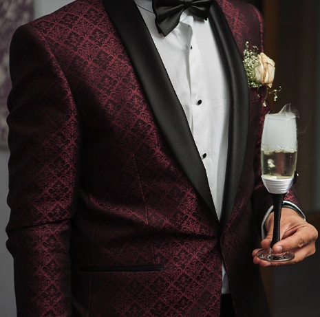 A man in a patterned burgundy suit with a black satin lapel holds a glass emitting smoke; he wears a bow tie, white shirt, and a boutonniere.