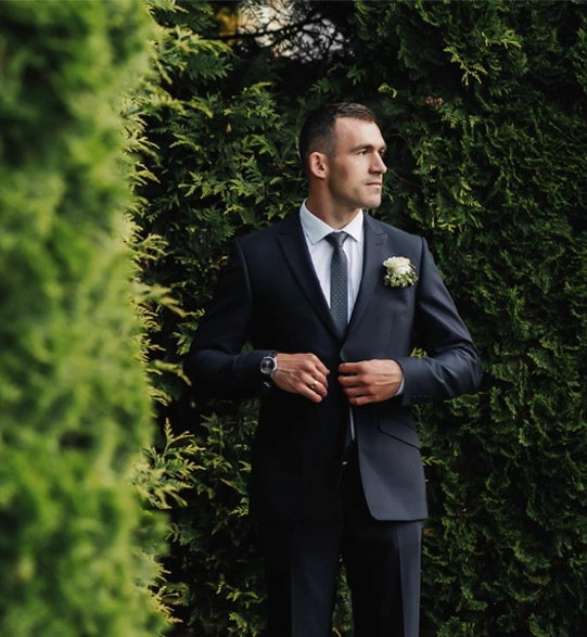 Man in a dark suit standing outdoors among green bushes, adjusting his jacket and looking to the side, with a boutonniere pinned to his lapel.