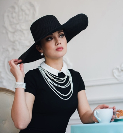 Woman wearing a black dress, pearl jewelry, and a wide-brimmed black hat sits at a table with a cup and a pastry in an elegant setting.