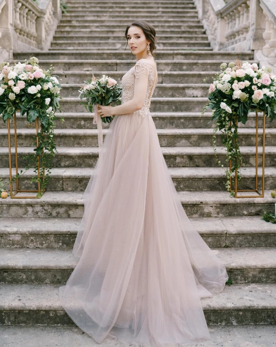 A woman in a light, flowing wedding dress stands on stone steps, holding a bouquet, with floral arrangements on both sides.