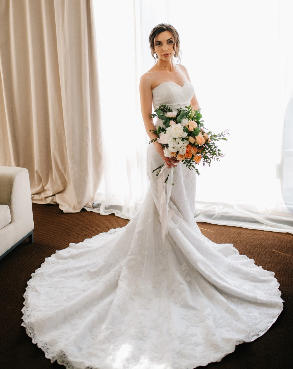 A bride in a strapless white wedding gown holds a bouquet of flowers, standing indoors in front of a light-filled window with beige curtains.