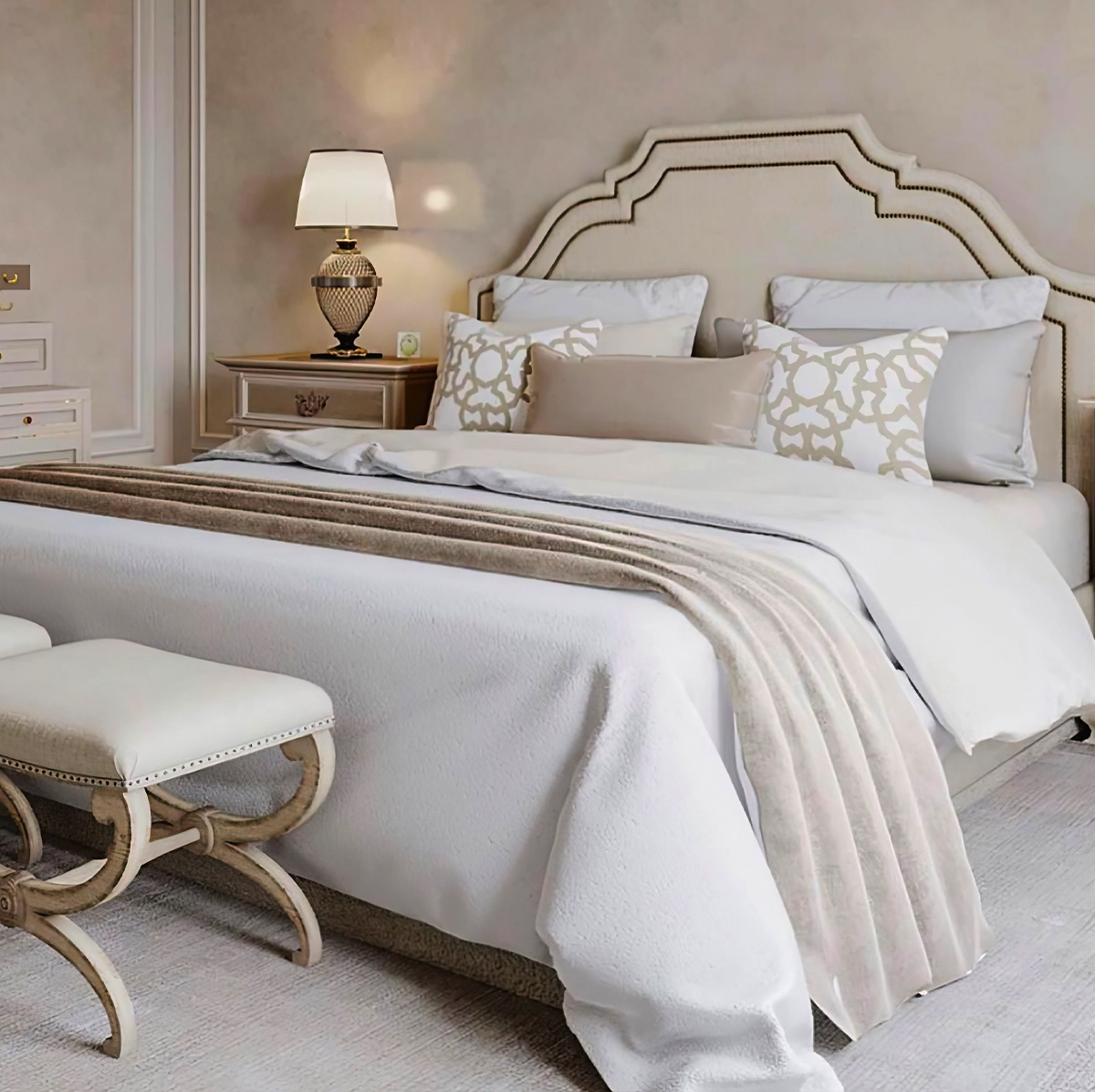 A neatly made bed with white and beige bedding, decorative pillows, a lamp on a nightstand, and an upholstered bench at the foot of the bed in a neutral-toned bedroom.
