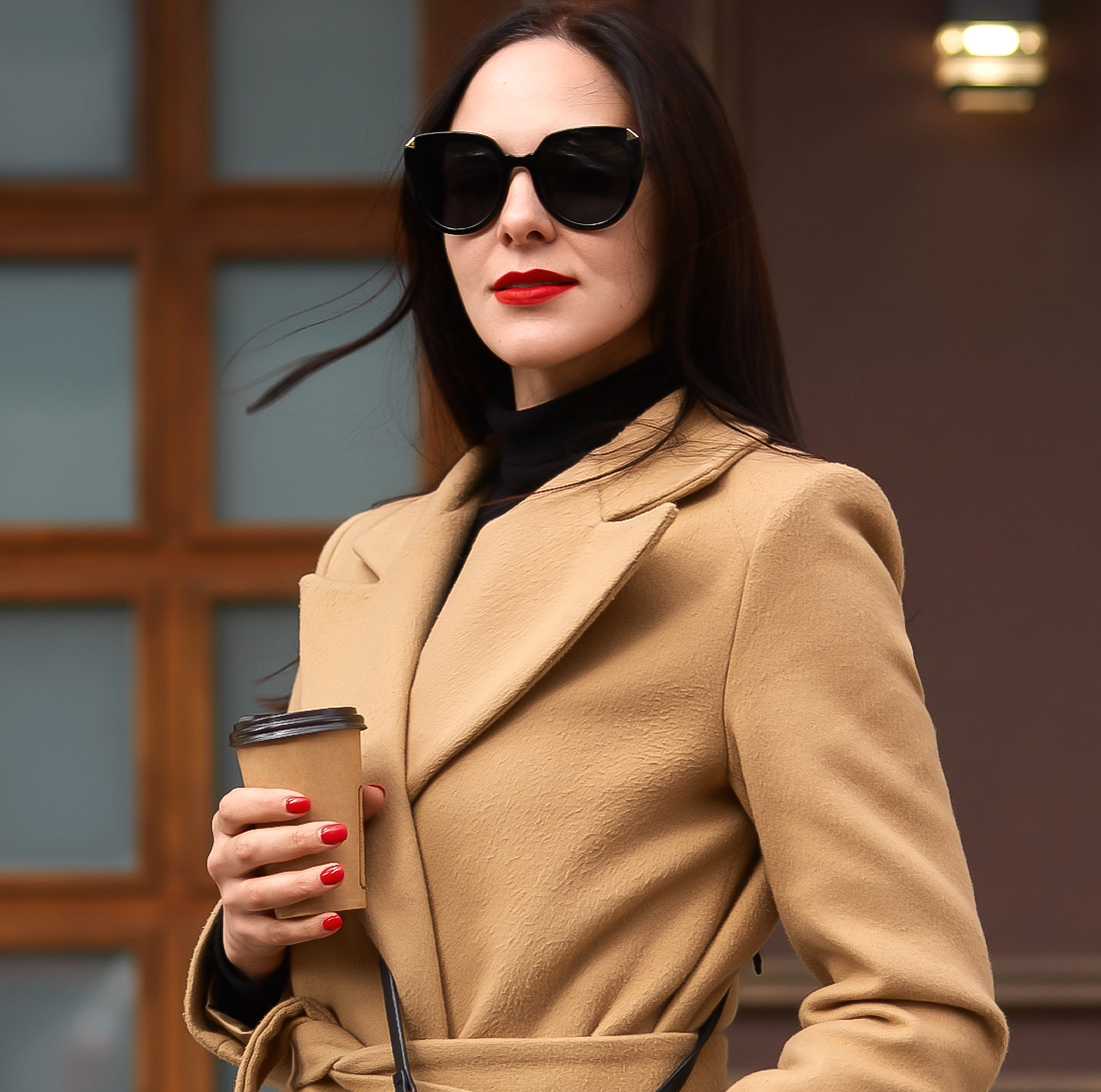 Woman in a tan coat and black sunglasses holds a coffee cup, standing outdoors in front of a building with wooden window frames.
