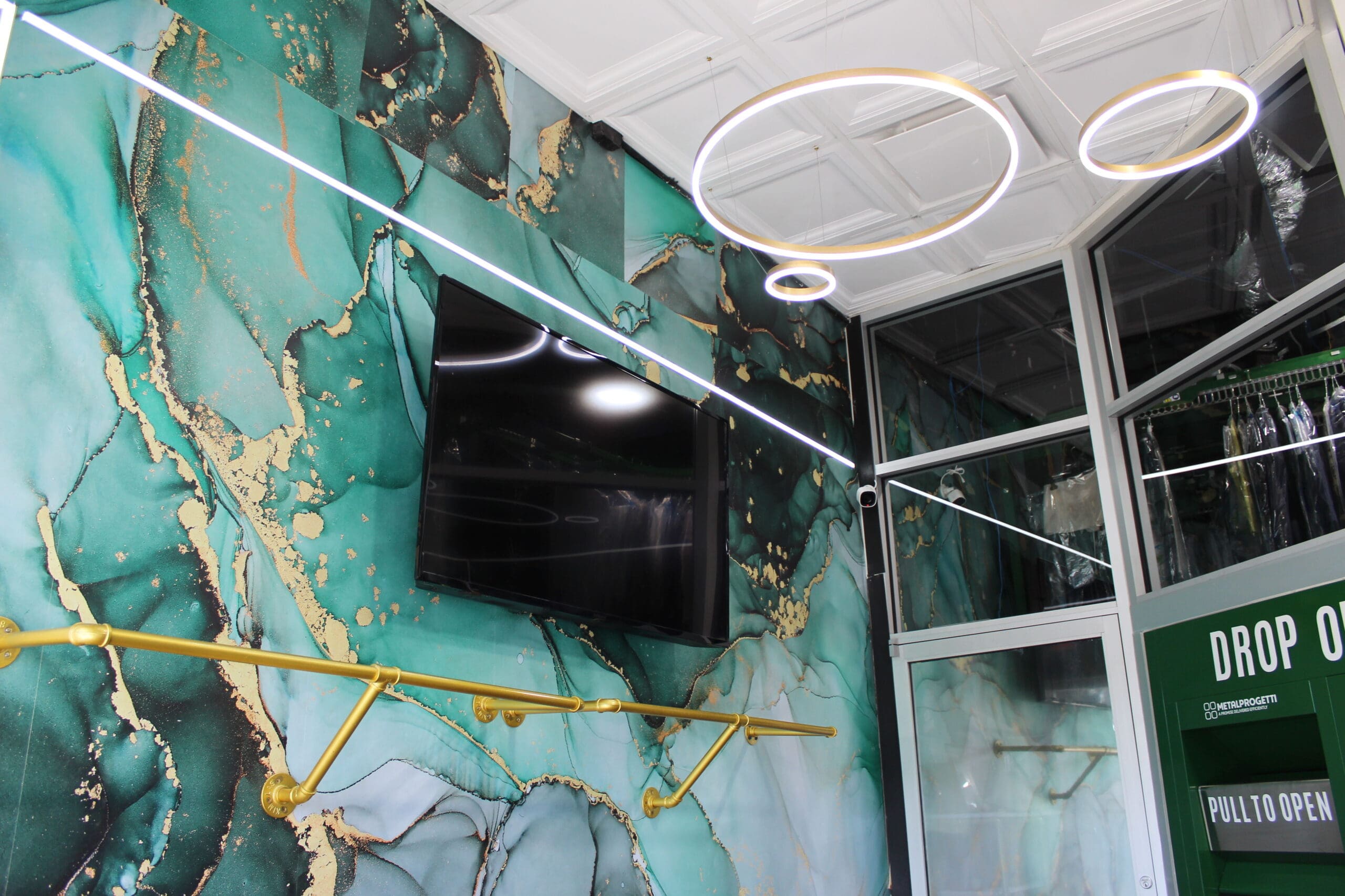 A modern interior with green and gold marble-patterned walls, a flat-screen TV, circular ceiling lights, and a "Drop Off" sign near a glass door.