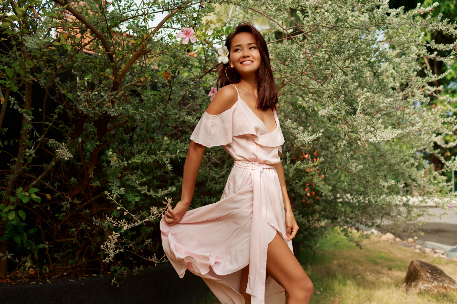 Woman in a light pink silk dress stands outdoors near green foliage, holding one side of her dress and smiling, showcasing the benefits of dry cleaning for delicate fabrics.
