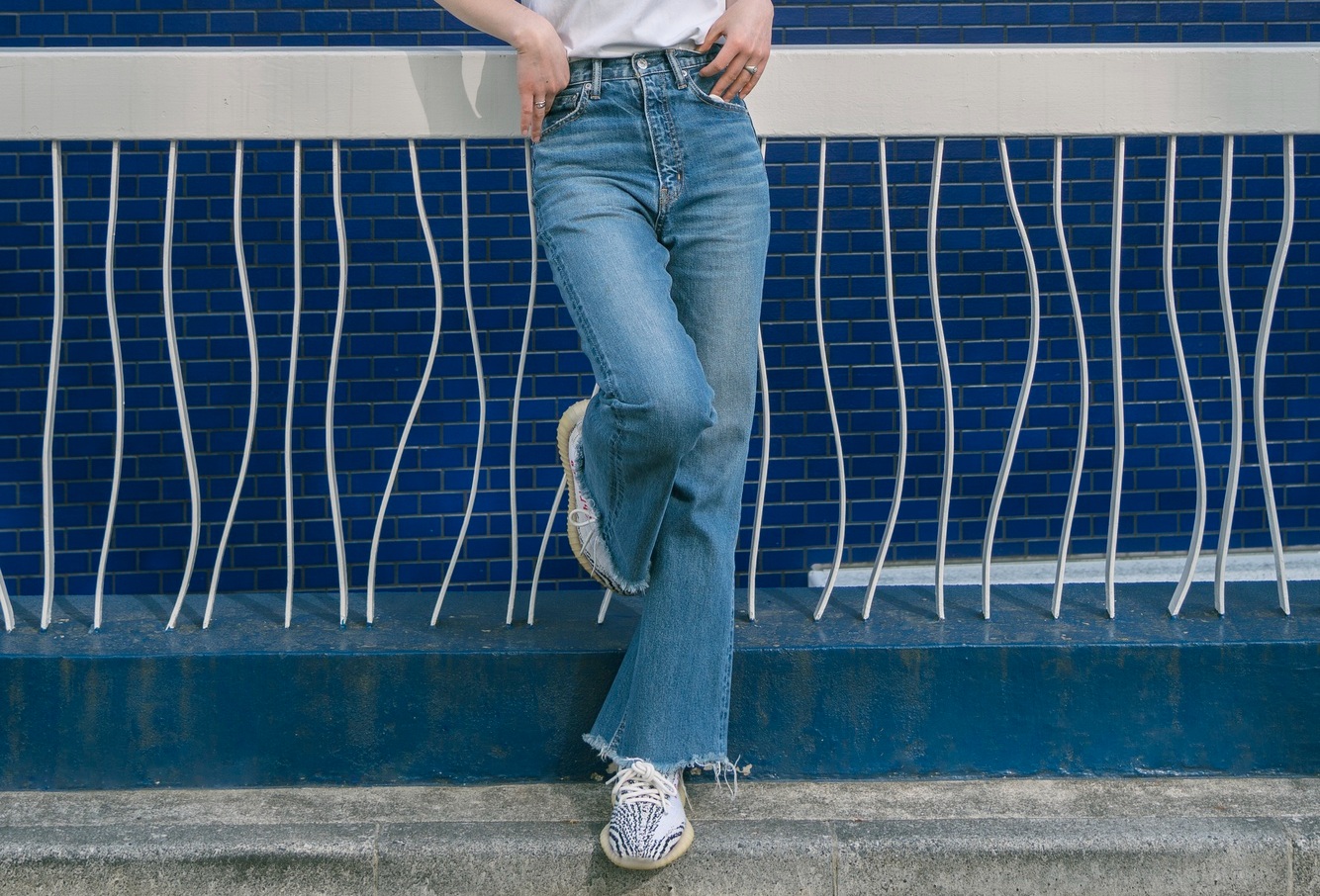 Person wearing blue jeans, white sneakers, and a white top standing against a blue tiled wall with white wavy bars; face out of frame.