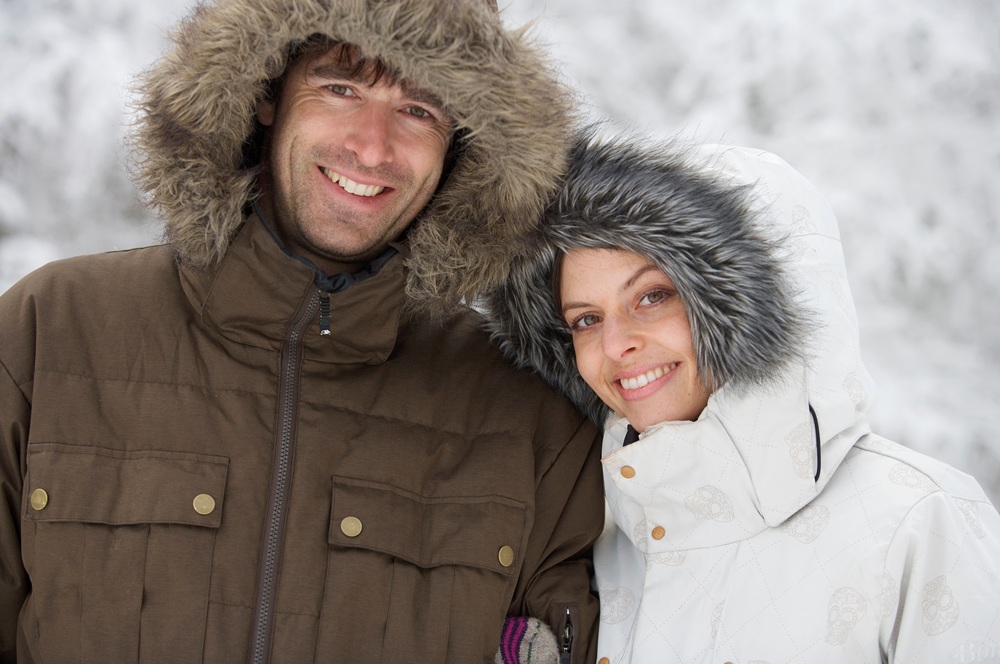 A man in a brown winter coat and a woman in a white winter coat, both with fur-lined hoods, standing together outdoors in a snowy setting.
