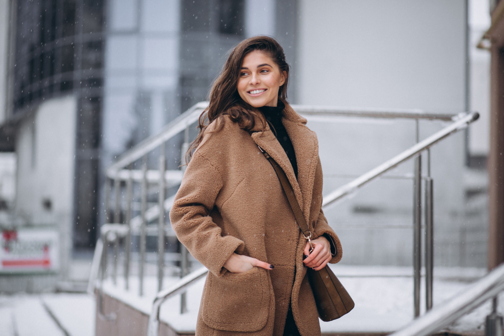 Woman wearing a brown coat stands outdoors on a snowy day, smiling and looking to the side, with a building and metal railings in the background.