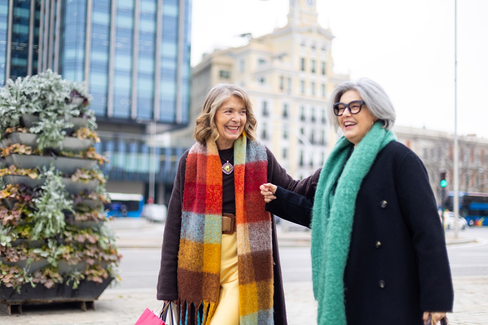 Two older women wearing colorful scarves and coats walk together outdoors in an urban area, smiling and holding hands.