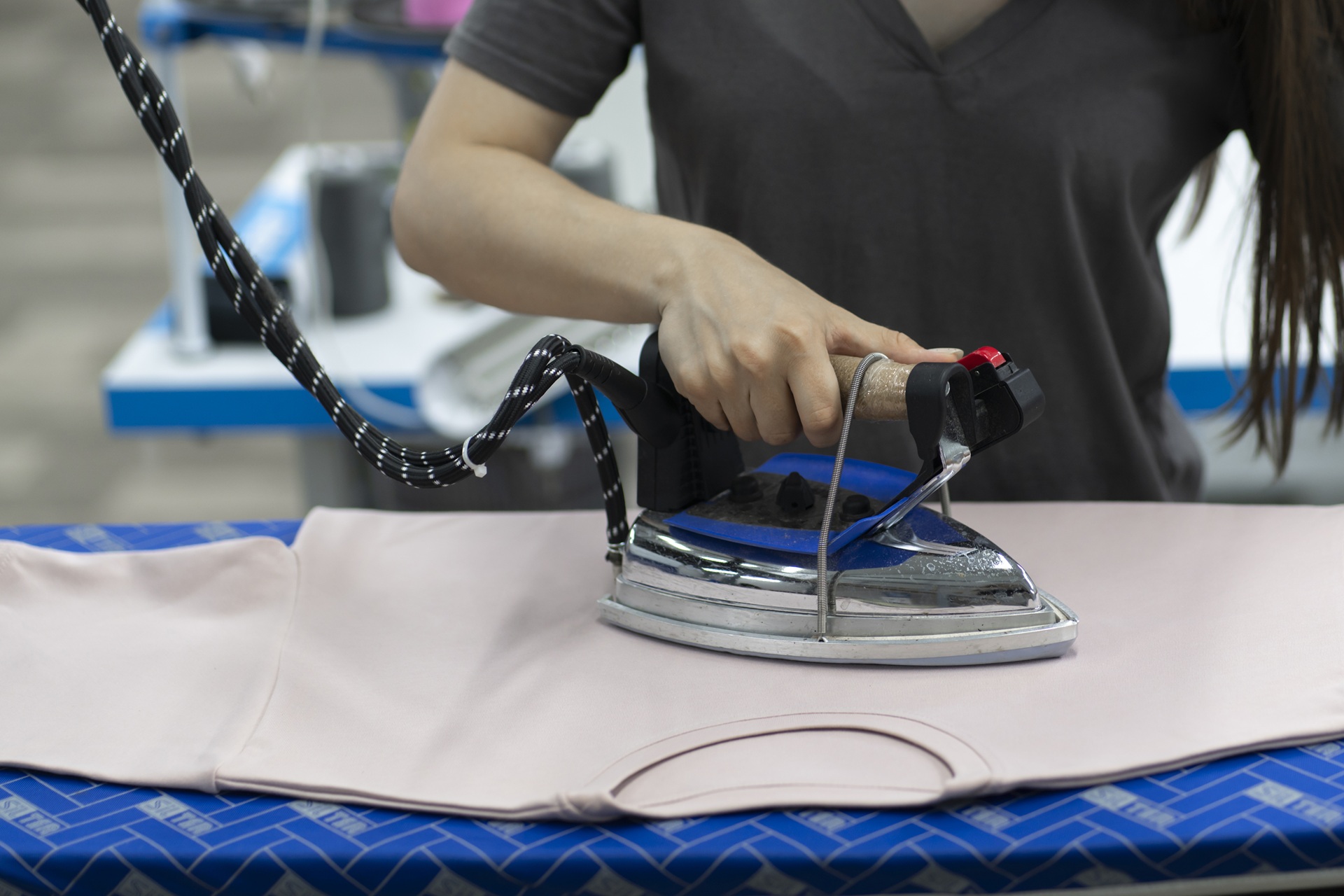 Person ironing a light pink t-shirt on a blue ironing board with a steam iron, with other sewing equipment visible in the background.