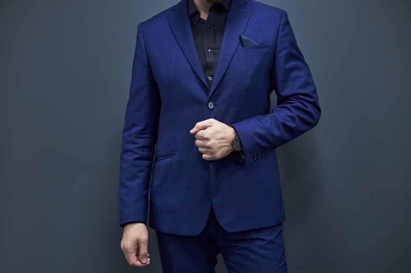 A man wearing a blue suit and black shirt stands against a dark background, with one hand adjusting his jacket.