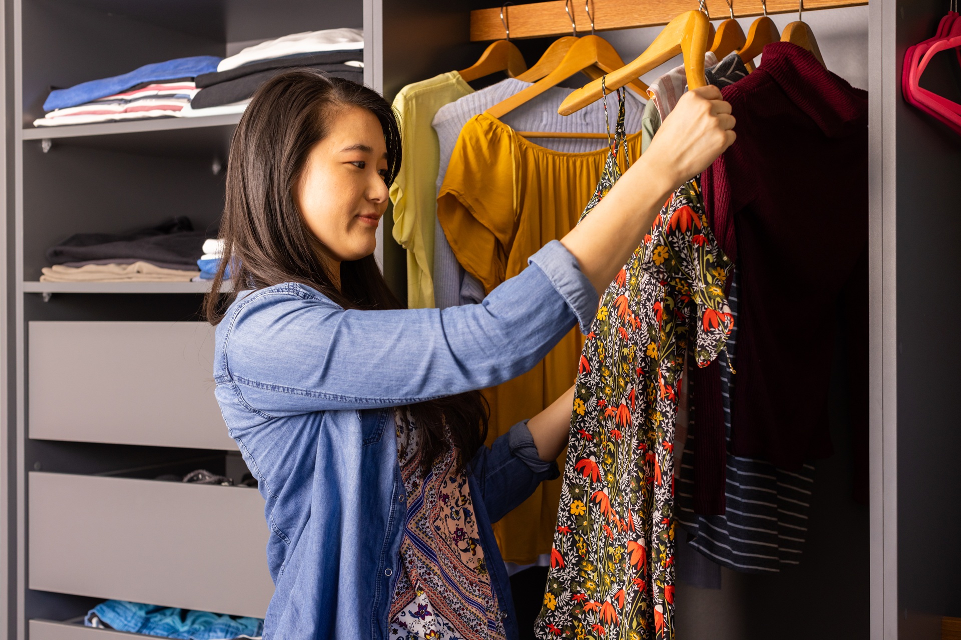 A woman stands in front of an open closet, holding up a floral dress and looking at it, with other clothes visible hanging and folded in the background.