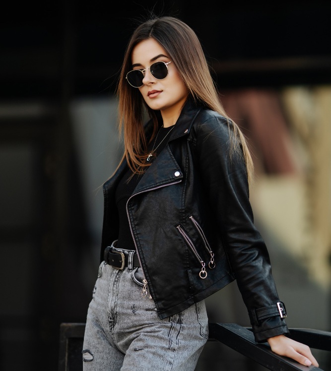 A woman wearing sunglasses, a black leather jacket, and gray jeans stands outdoors, resting one arm on a railing.