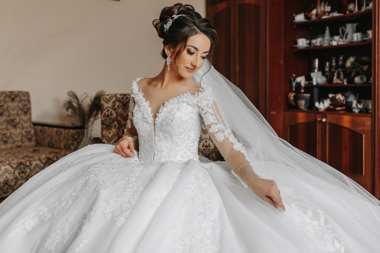 A bride in an ornate white wedding dress with lace details and a veil sits indoors, looking down and touching her gown—reminding us how soon you should preserve a wedding dress to keep its beauty for years to come.